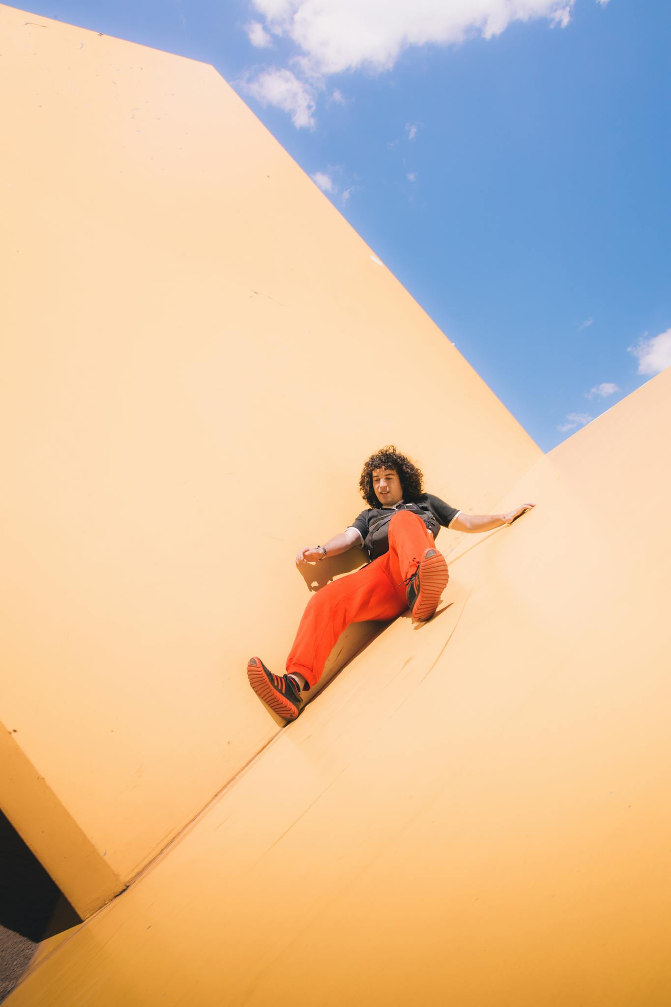 A young man in red pants sits on a large yellow geometric wall against a clear blue sky.